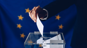 A hand places a ballot paper into a transparent ballot box with a blue background and yellow stars.