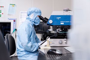 Laboratory worker wearing a full blue protective gown seated at a desk using a microscope.