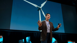 Photograph of a woman making a presentation to an unseen audience, standing in front of a large screen showing a wind turbine, with a microphone in her hand.