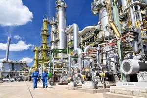 Workers in work clothes and hard hats walk outside on a sunny day in a refinery plant with huge metal pipes and machinery.