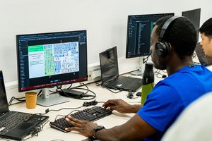 African man wearing headphones, looking at a screen, typing on a keyboard in a working environment