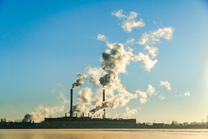 Thick smoke rising from two tall industrial chimneys and spreading across  the blue skies above.