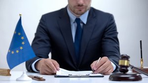 Photograph of a suited man correcting a document, seated beside a blue European flag and a gavel.