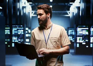 Photograph of a bearded man, holding a laptop, in a room full of data servers.