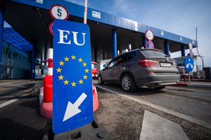 EU border crossing point with a blue EU sign displaying the circle of yellow stars and a white directional arrow in the foreground, while a car approaches the checkpoint booths in the background.