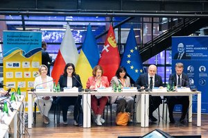Panel of speakers at the European Alliance of Cities and Regions for the Reconstruction of Ukraine – political level meeting Gdańsk - Poland - June 2026. Behind them the Polish, Ukrainian, EU & Gdańsk flags.