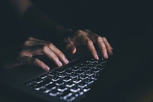 Hands typing on a laptop keyboard in dim lighting, suggesting potentially illicit online activity.