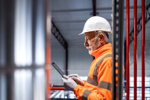 Bearded man with white helmet, grey gloves and orange jacket checks his ipad in an industrial building.