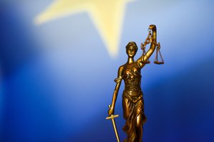 Photograph of a bronze statuette of Themis, goddess of divine justice, with the EU flag as a backdrop.
