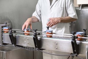 Worker in white protective clothing checking jars on a stainless-steel conveyor belt in a production facility.