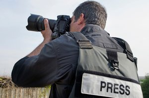 A photojournalist wearing a press vest and taking a photograph outdoors.