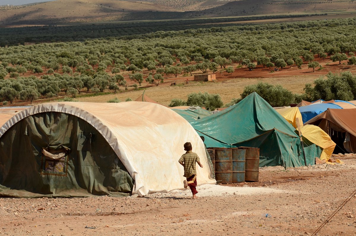 Tents lined up in a refugee camp, with trees and open plains in the background and a barefoot child running alongside.