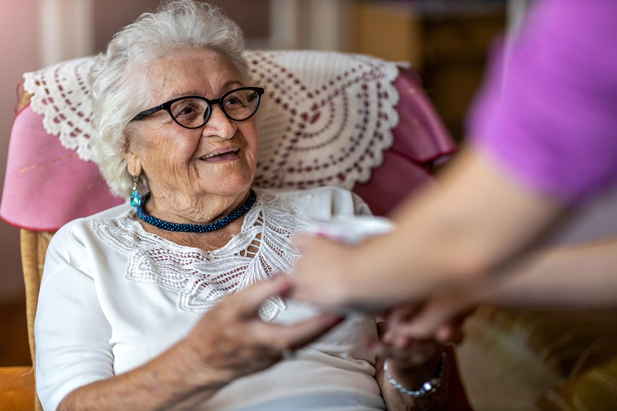 A nurse handing something to a smiling elderly lady sitting on a chair.