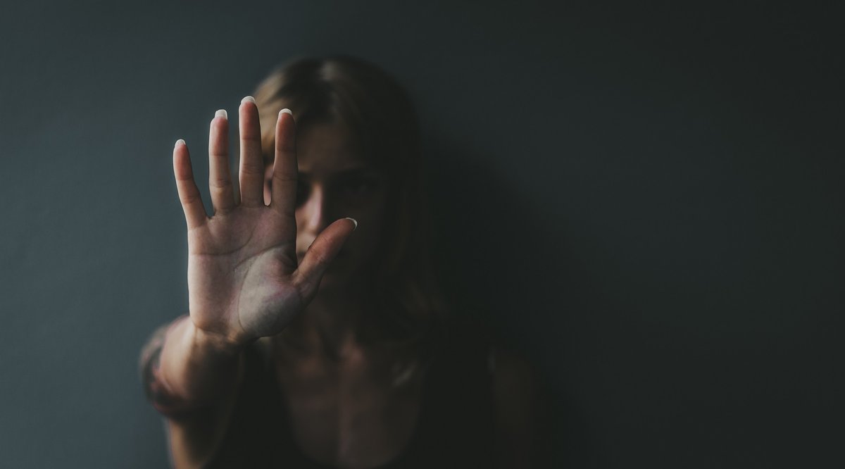 A woman is shown standing against the background of a dark wall with her hand raised in a gesture of self-defence.
