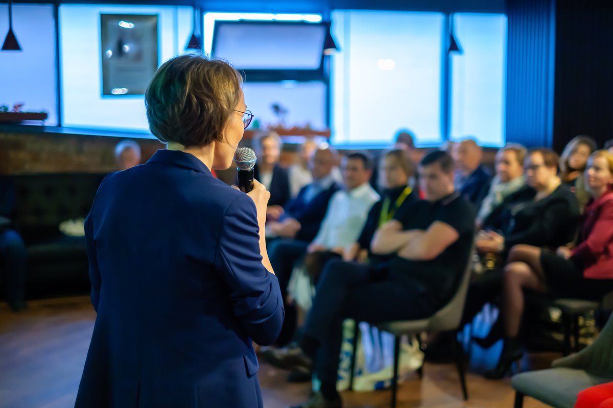 Photograph of a woman, seen from behind, speaking to a seated audience.