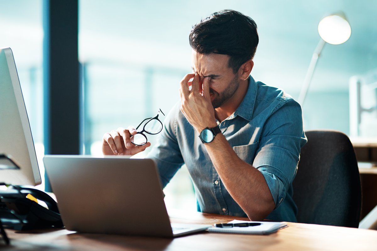Photo of a male employee sitting&nbsp;at a desk,&nbsp;rubbing his face, indicating possible mental fatigue and emotional strain