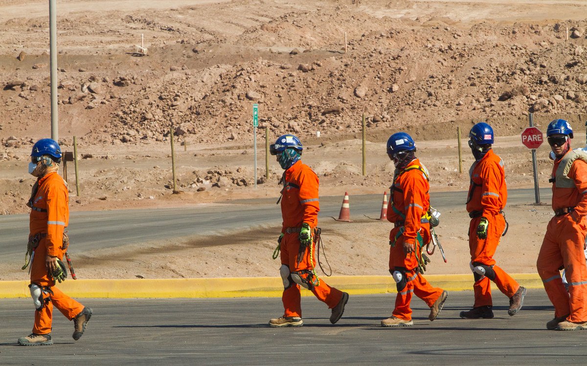 Workers in protective clothing walking across a road at an industrial mining site in an arid landscape.
