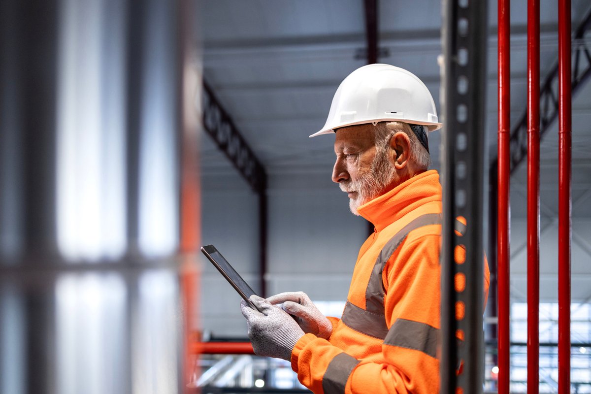 Bearded man with white helmet, grey gloves and orange jacket checks his ipad in an industrial building.