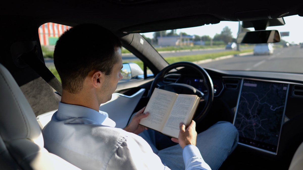 Photograph of a man reading a book behind the steering wheel of a self-driving car.