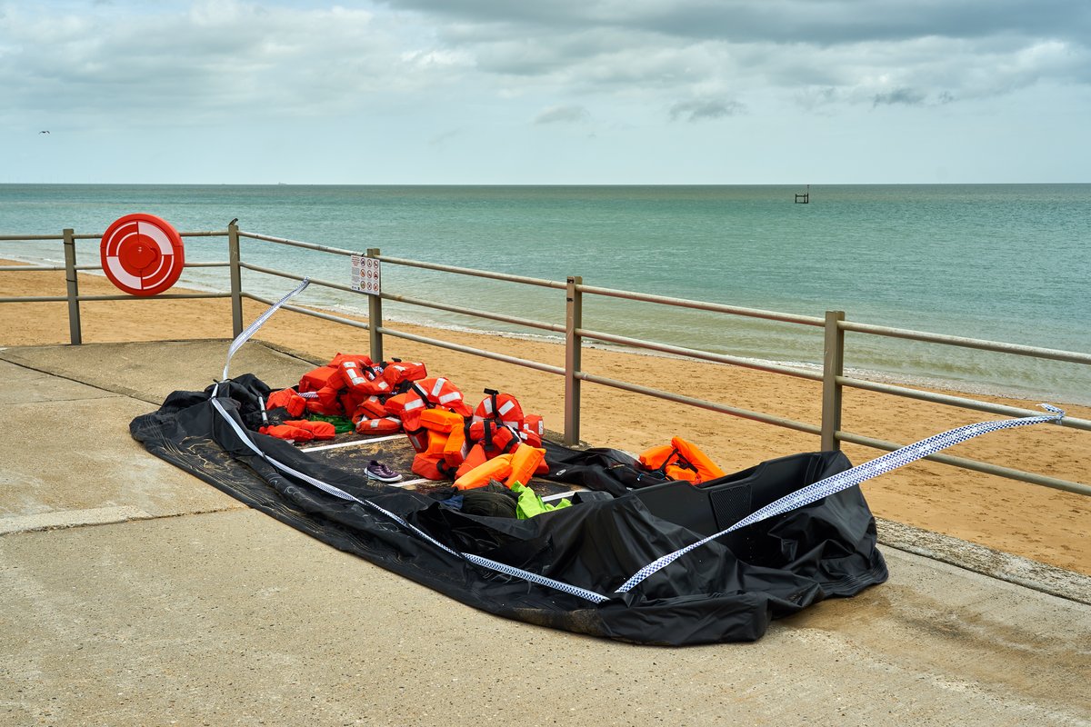 The image shows a deflated black dinghy filled with life vests left on a seafront promenade behind barrier tape.