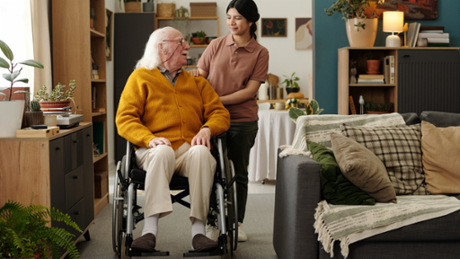 Photograph of an elderly man in wheelchair talking to a young Latin female carer in a living room