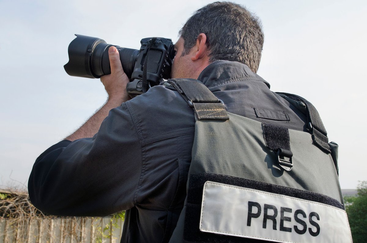 A photojournalist wearing a press vest and taking a photograph outdoors.